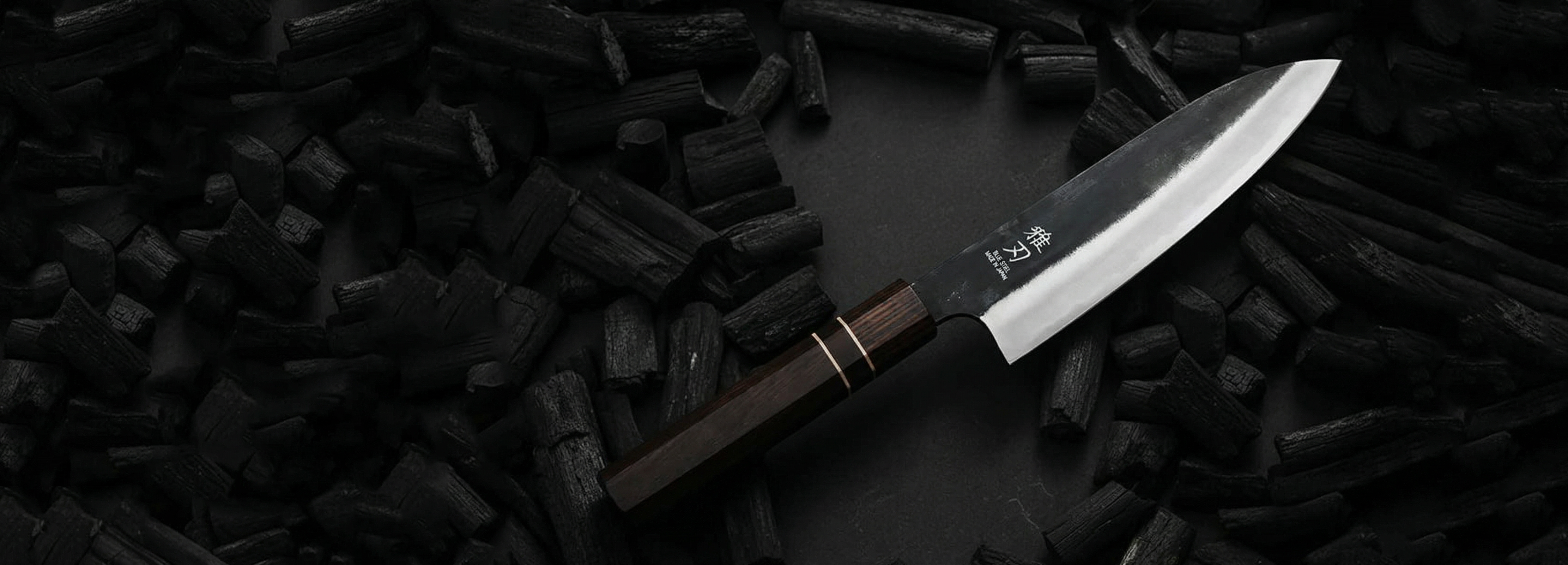 A professional Japanese chef's knife with a black-forged blade and dark wood handle, resting on a bed of natural black charcoal chunks